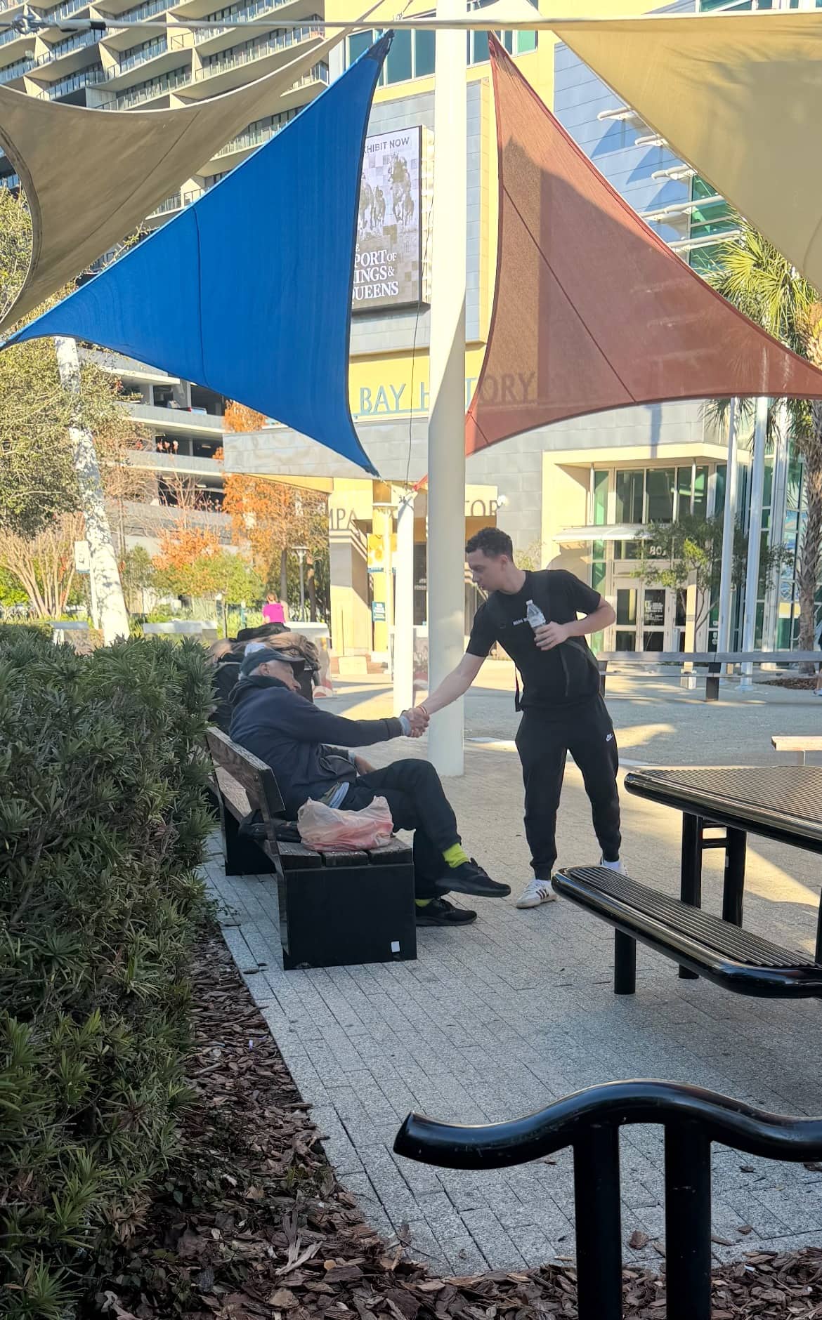A Making Heaven Crowded volunteer holding a water bottle shakes hands with a man seated on a bench in a downtown Tampa plaza