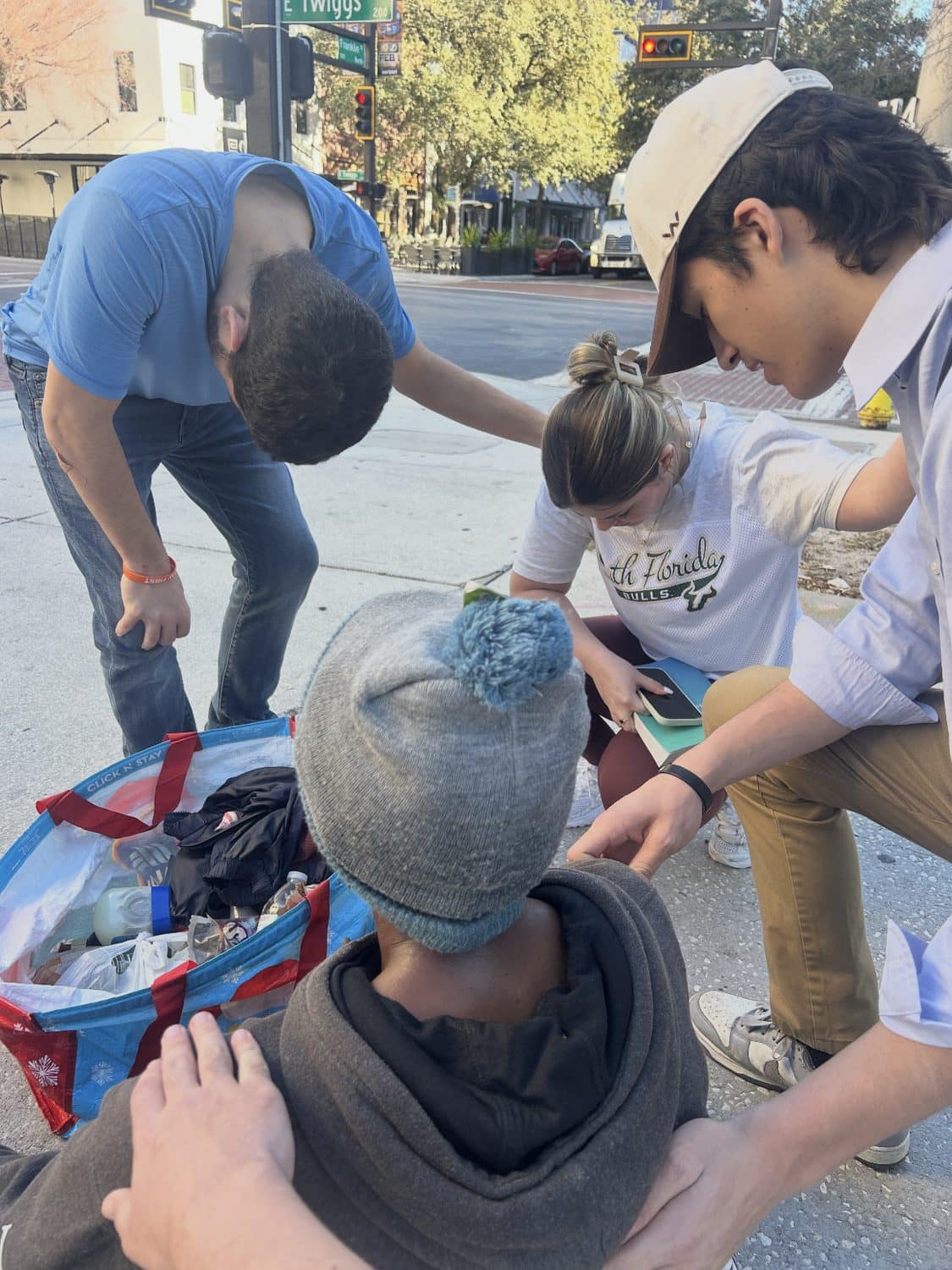 Volunteers kneeling with a man on a downtown Tampa sidewalk, one of them holding his shoulder